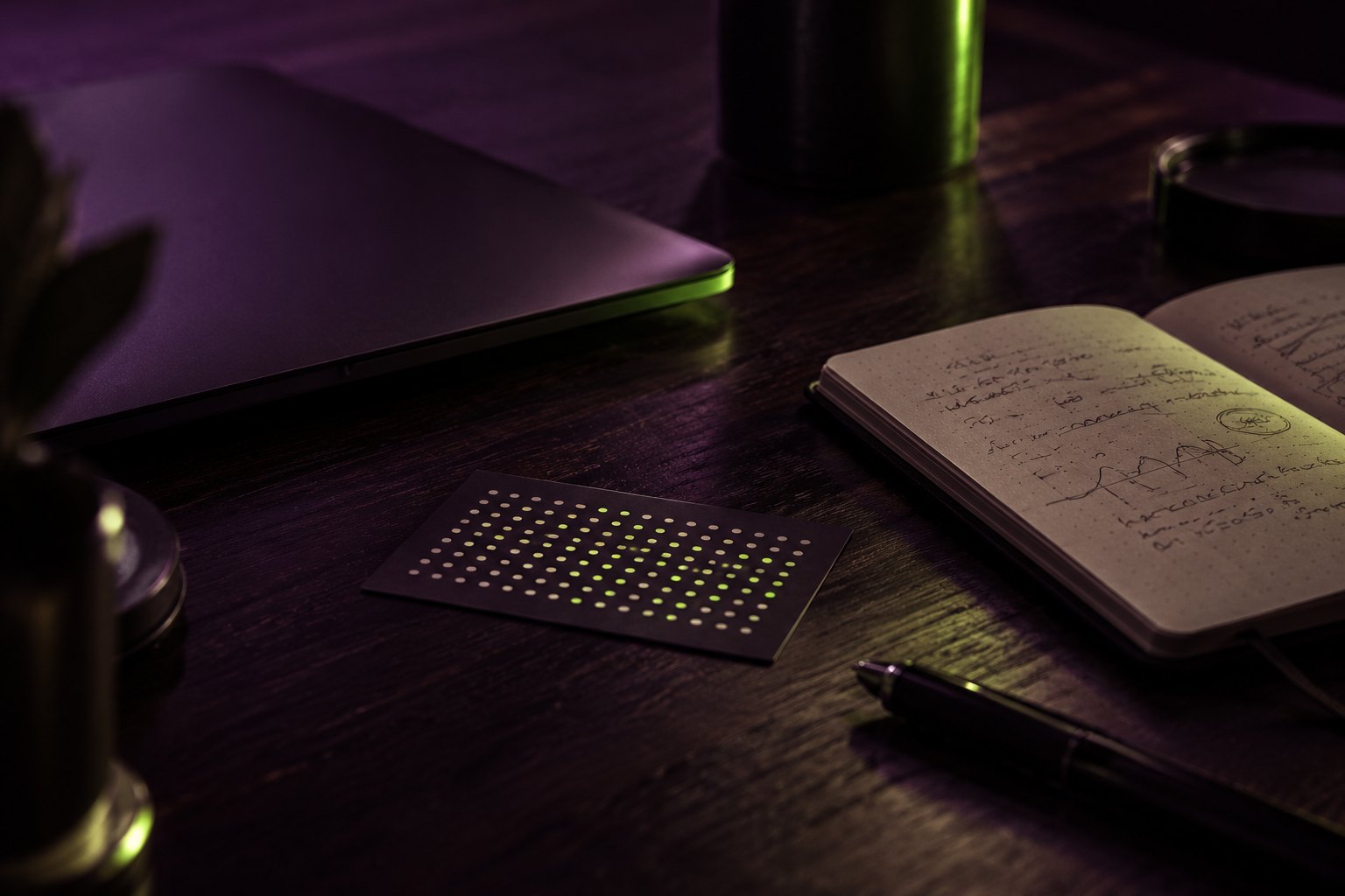 A dark desk at night: a closed laptop, a notebook full of handwritten notes and sketches, and a small card printed with a grid of dots, some glowing green
